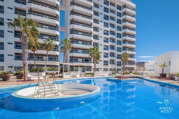 A hotel pool area with a circular hot tub, lounge chairs, and palm trees.