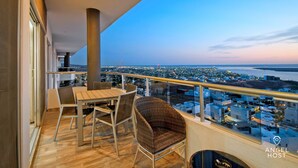 Un balcón con una mesa y sillas de madera, una silla de mimbre y vistas a un paisaje urbano al atardecer.