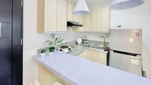 Cocina moderna con armarios de madera clara, encimera blanca, horno empotrado, refrigerador de acero inoxidable e iluminación colgante.