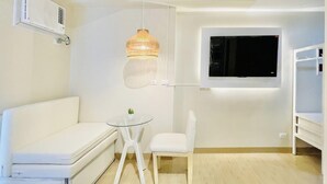 Une pièce moderne et minimaliste, avec un canapé blanc, une petite table ronde agrémentée d’une plante, un luminaire suspendu et un téléviseur fixé au mur.