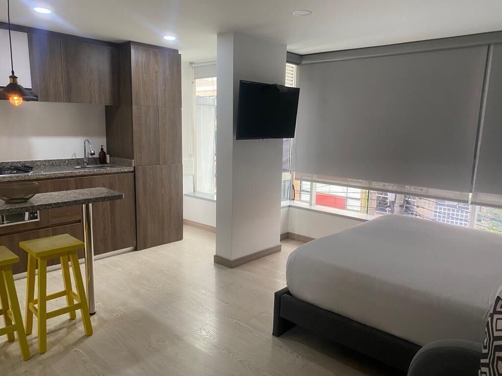 A modern room with a bed, a desk with stools, a TV, and a window with blinds.