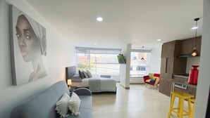 A modern living room with a grey sofa, a dining area with a yellow stool, and a kitchen with wooden cabinets. - Spacio30 (Bogotá)