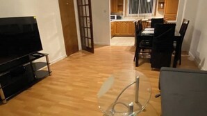Un salon avec un téléviseur, une table en verre et un coin repas avec des chaises, et une cuisine en arrière-plan.