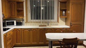 Une cuisine avec des meubles de rangement en bois, un plan de travail blanc, un four à micro-ondes et une fenêtre avec des stores.