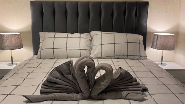 Un lit doté d’une tête de lit capitonnée, de deux lampes de chevet et d’une serviette disposée en forme de cœur sur le lit.