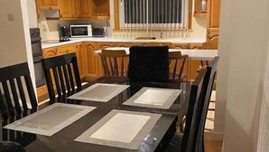 Un coin repas avec une table et des chaises en bois foncé, une cuisine à l’arrière-plan et une fenêtre avec des stores.