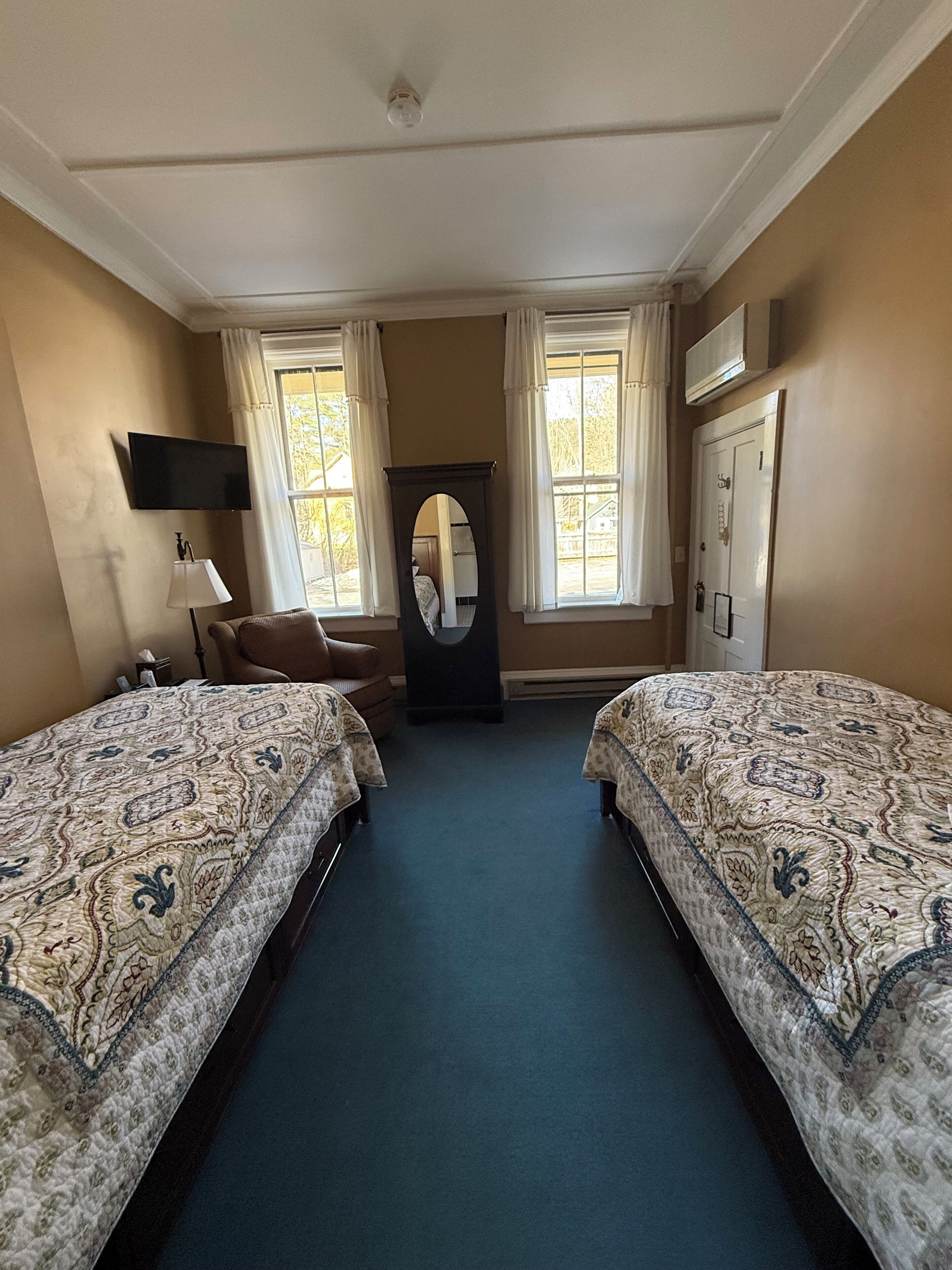 A hotel room with two beds, a chair, a mirror, a TV, and a door.