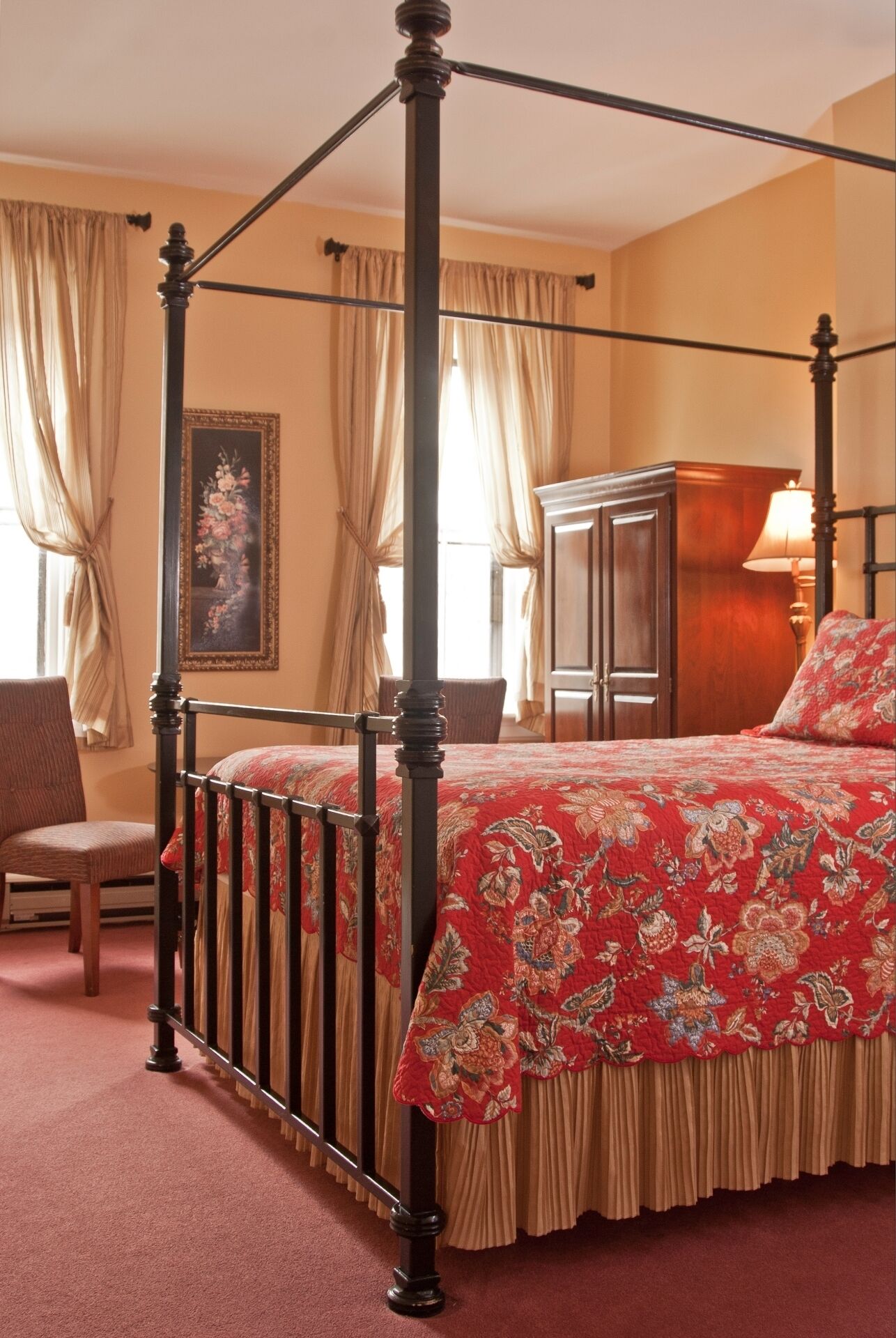 Una cama con dosel con colcha estampada, una mesita de noche de madera, una silla y un cuadro en la pared.