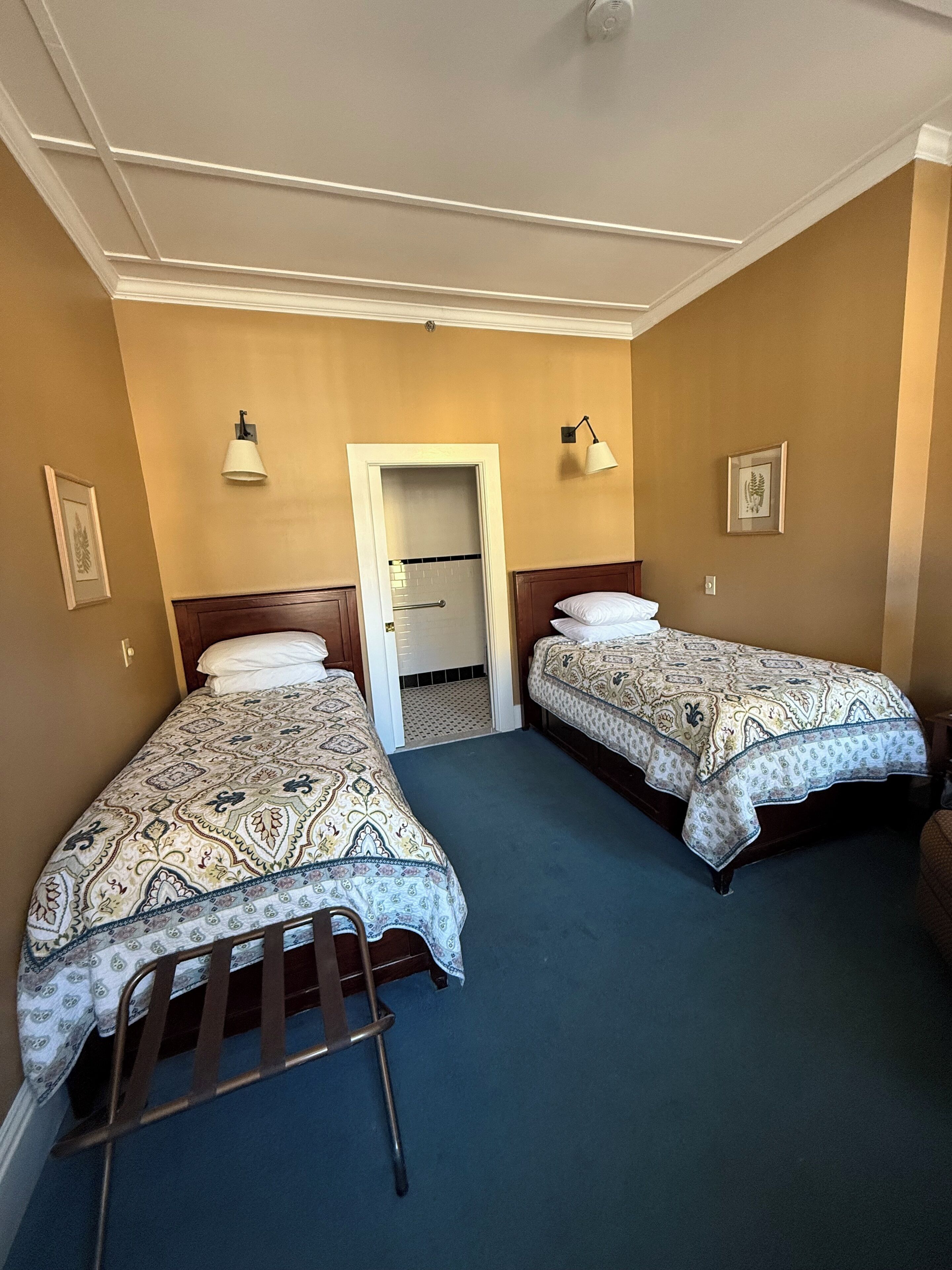 Habitación de hotel con dos camas individuales, cabecero de madera, alfombra azul y una puerta blanca que da al baño.