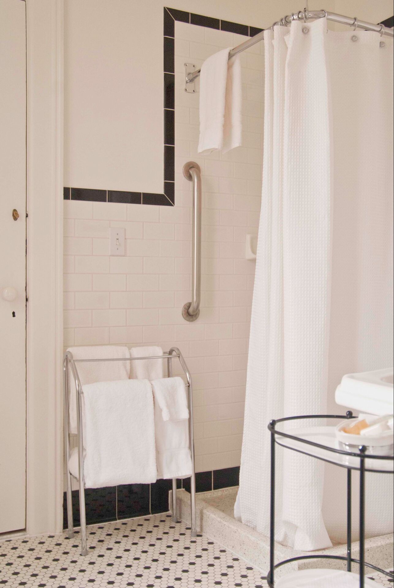 A bathroom with a white tiled floor, a shower with a white curtain, a towel rack with two towels, and a small table with a soap dish.