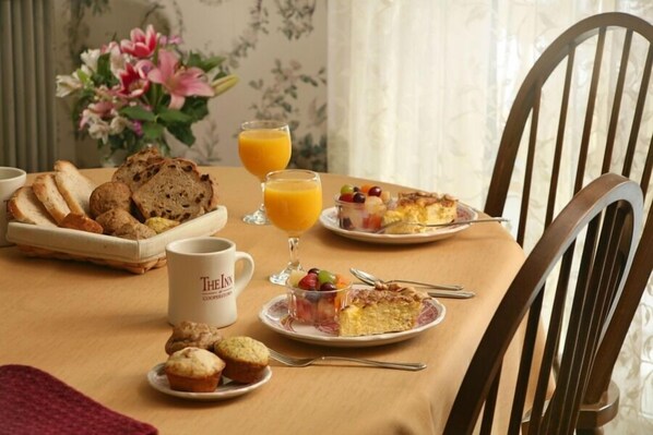 A breakfast table with bread, muffins, a muffin, a glass of orange juice, and a plate with a slice of pie.