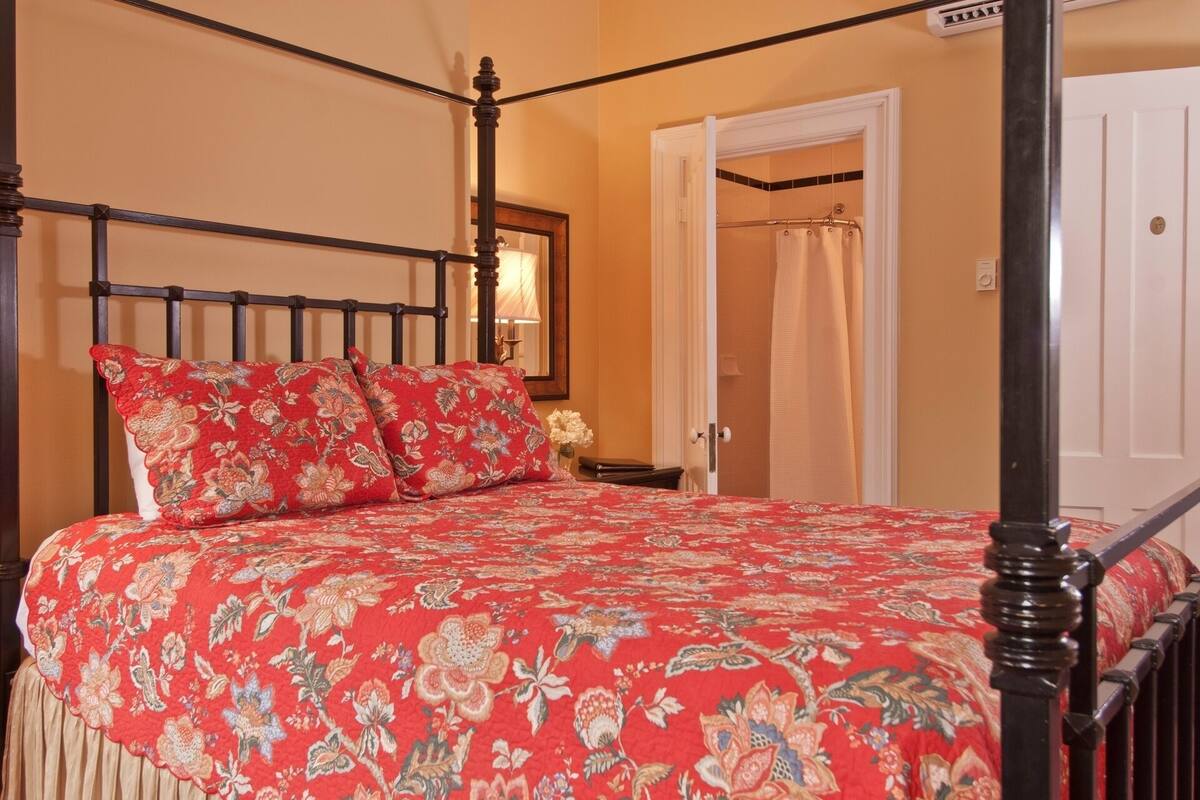 A four-poster bed with a floral bedspread, a nightstand with a lamp, and a door leading to a bathroom.