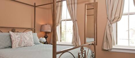 A four-poster bed with a quilted bedspread, a bedside table with a lamp, and a large mirror on the wall.