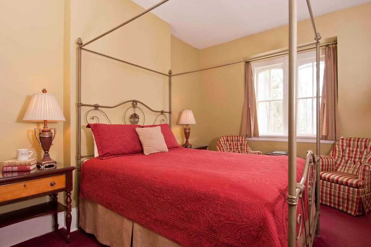 A four-poster bed with a red bedspread, two bedside lamps, a wooden nightstand, a plaid armchair, and a window with curtains.