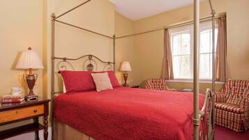 A four-poster bed with a red bedspread, two bedside lamps, a wooden nightstand, a plaid armchair, and a window with curtains.