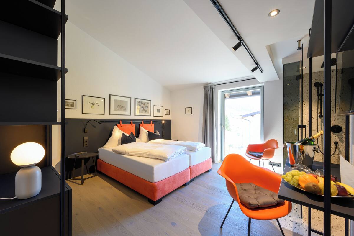 A modern hotel room with a bed, a desk with a lamp, a chair, and a breakfast bar with fruit.