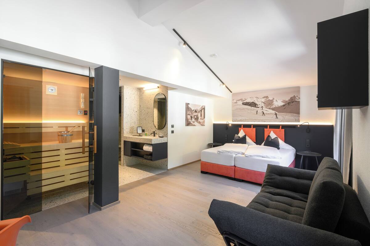 A modern hotel room with a bed, a sofa, a TV, a bathroom with a mirror, and a picture of a mountain scene on the wall.