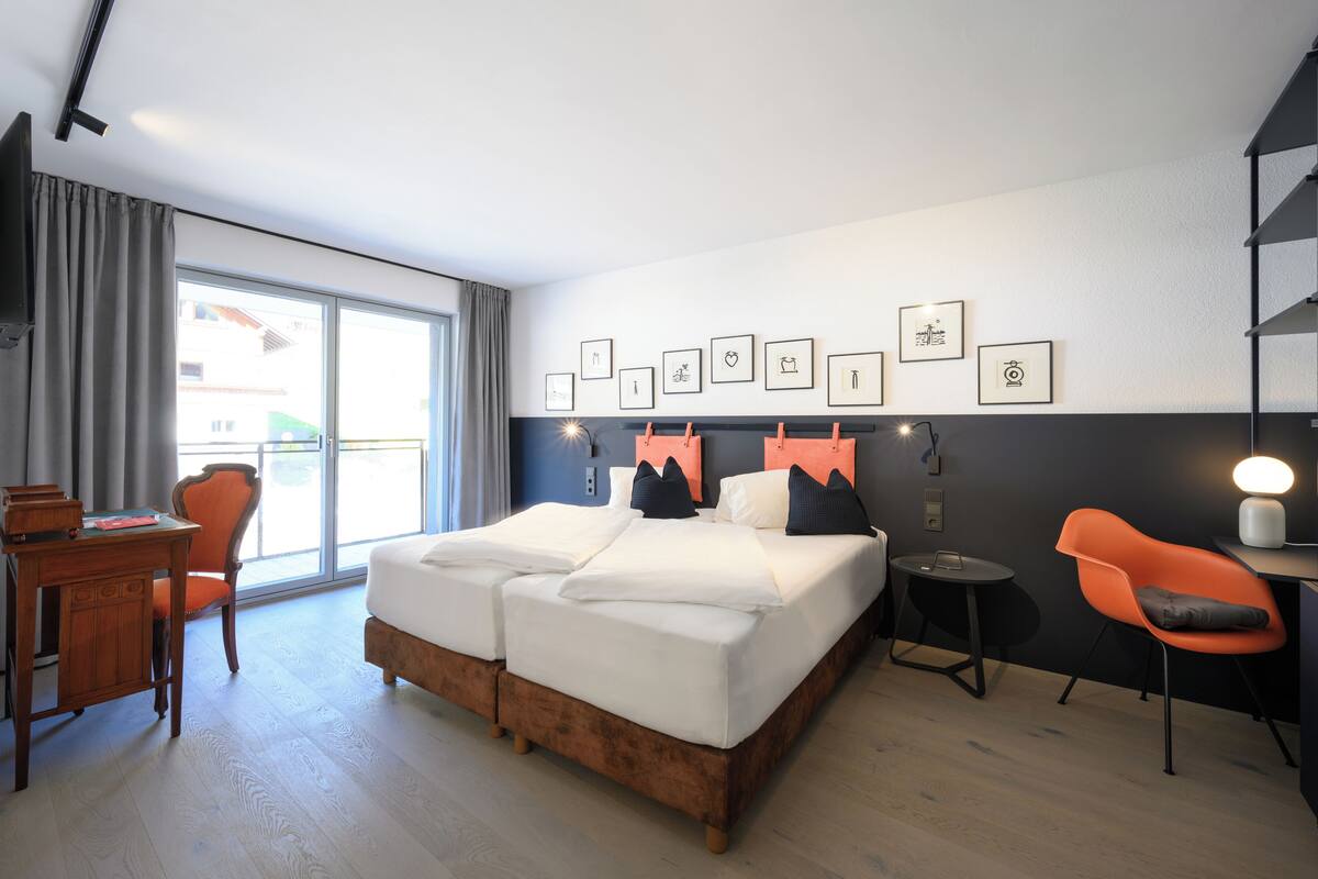 A modern hotel room with a large bed, a desk, and a chair. There are framed pictures on the wall and a window with curtains.