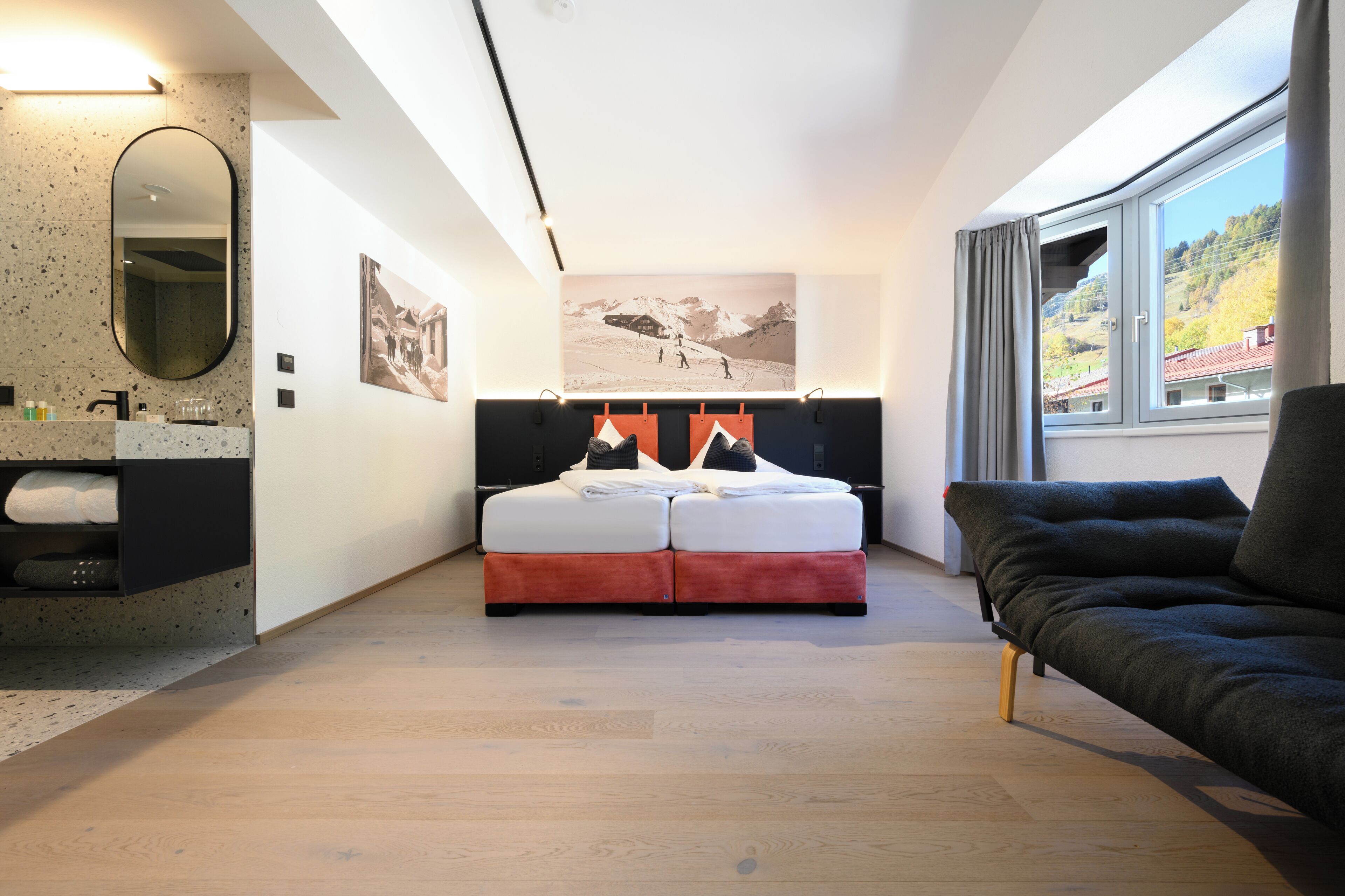 Een moderne hotelkamer met een bed, een sofa, een badkamer met spiegel en wastafel, en een groot raam met uitzicht op bergen.