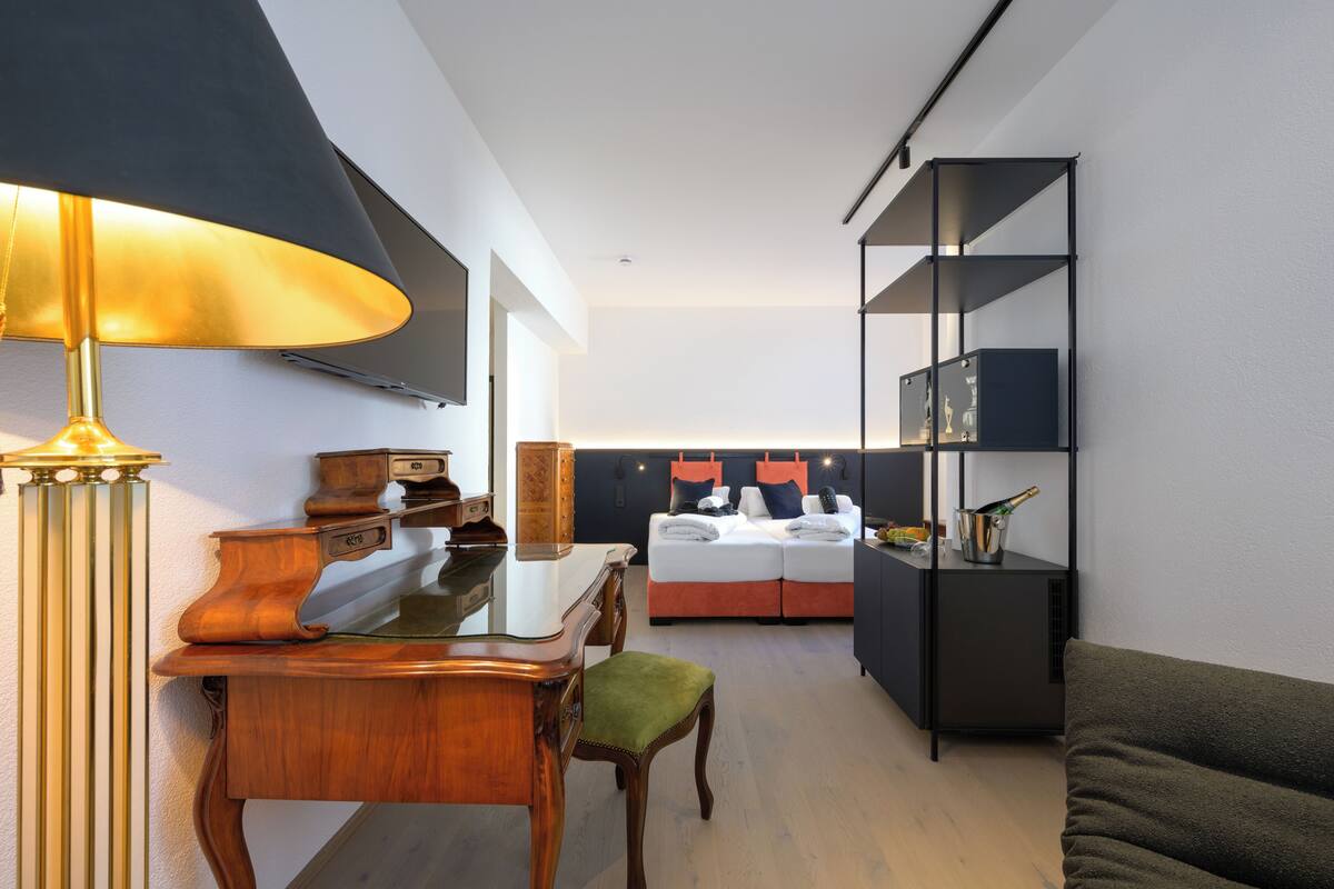 A modern living room with a wooden desk, a lamp, a sofa, a bed with orange pillows, and a black shelving unit.