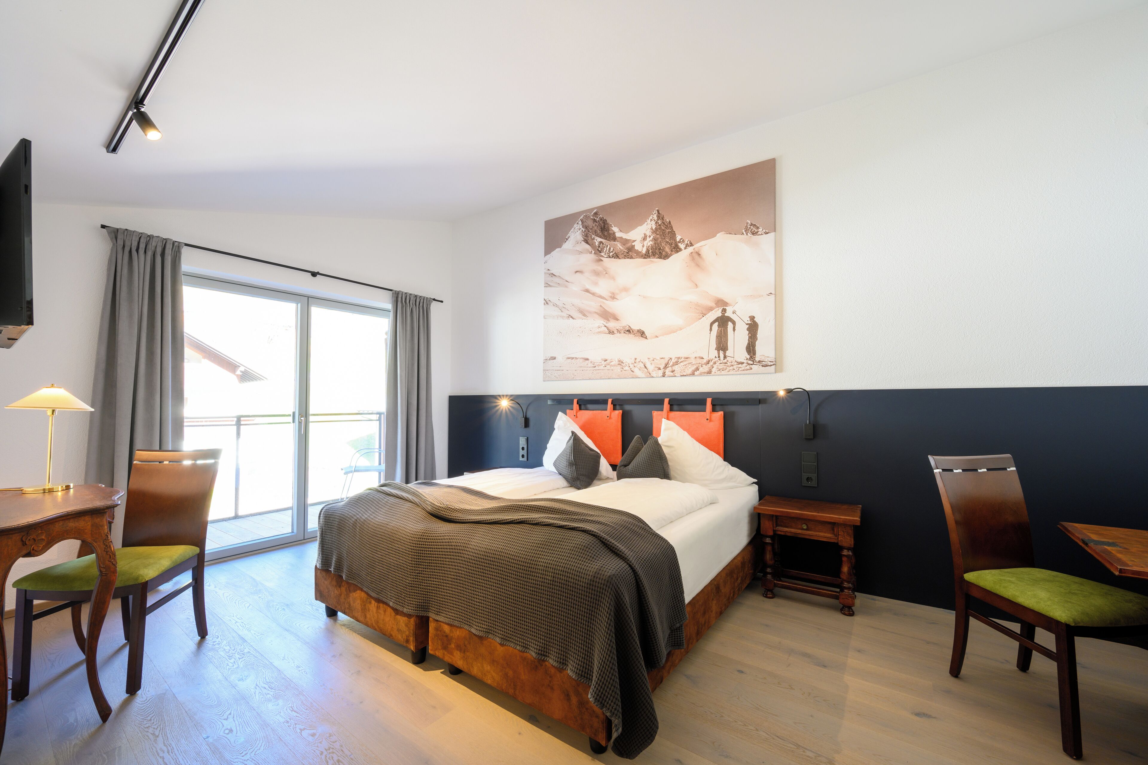 A hotel room with a large bed, a desk, two chairs, a window with curtains, and a wall-mounted painting.