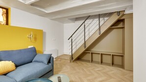 A modern living room with a blue sofa, a glass coffee table, and a wooden herringbone floor. A staircase with a metal railing leads to an upper level. - TBS 51 - Spacious and modern duplex apartment (Paris)