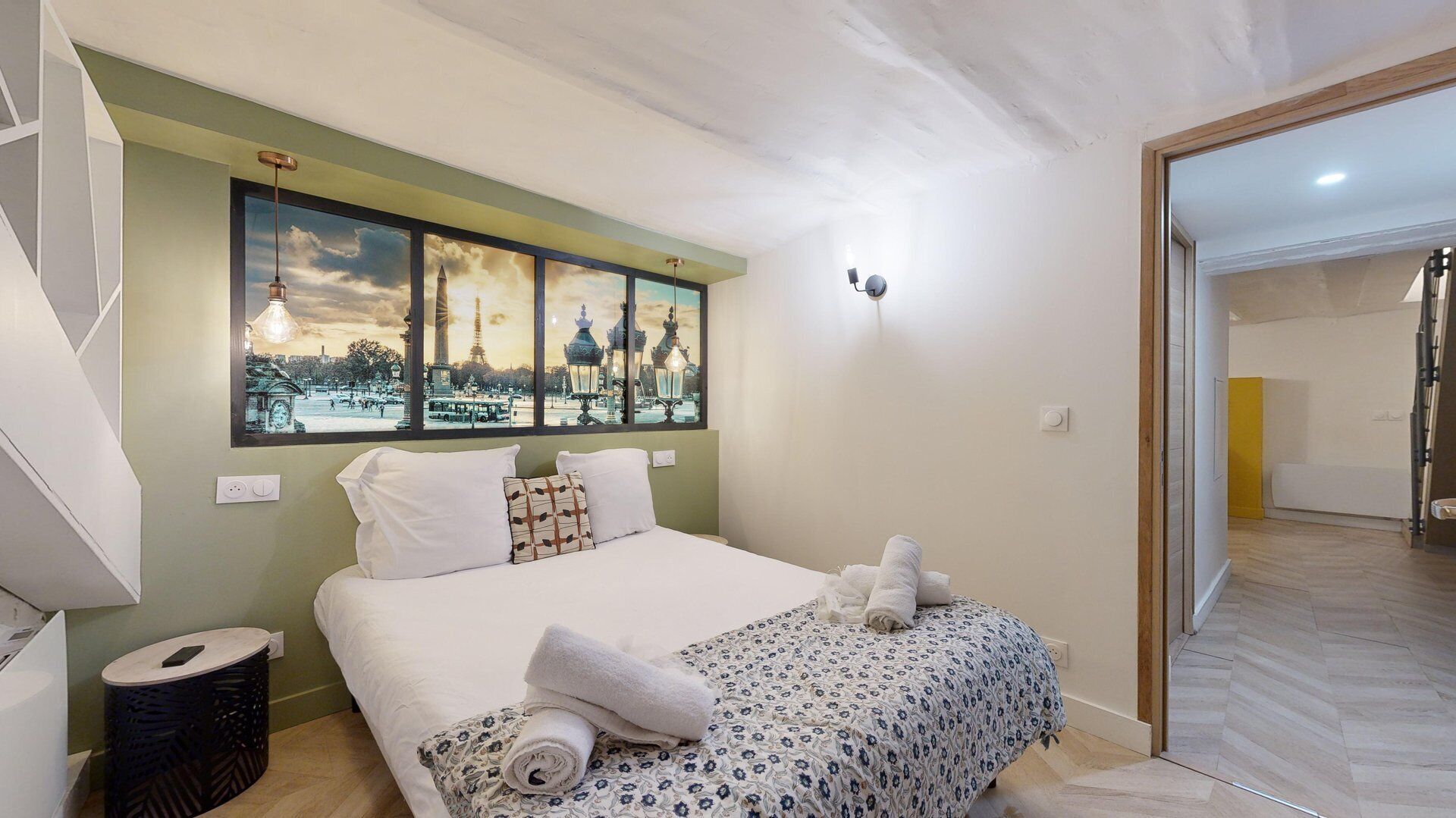 Un dormitorio con cama, una ventana con vista a la ciudad, una lámpara de noche y una mesita redonda.