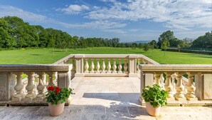 A balcony with a stone balustrade overlooking a well-manicured lawn and distant trees.