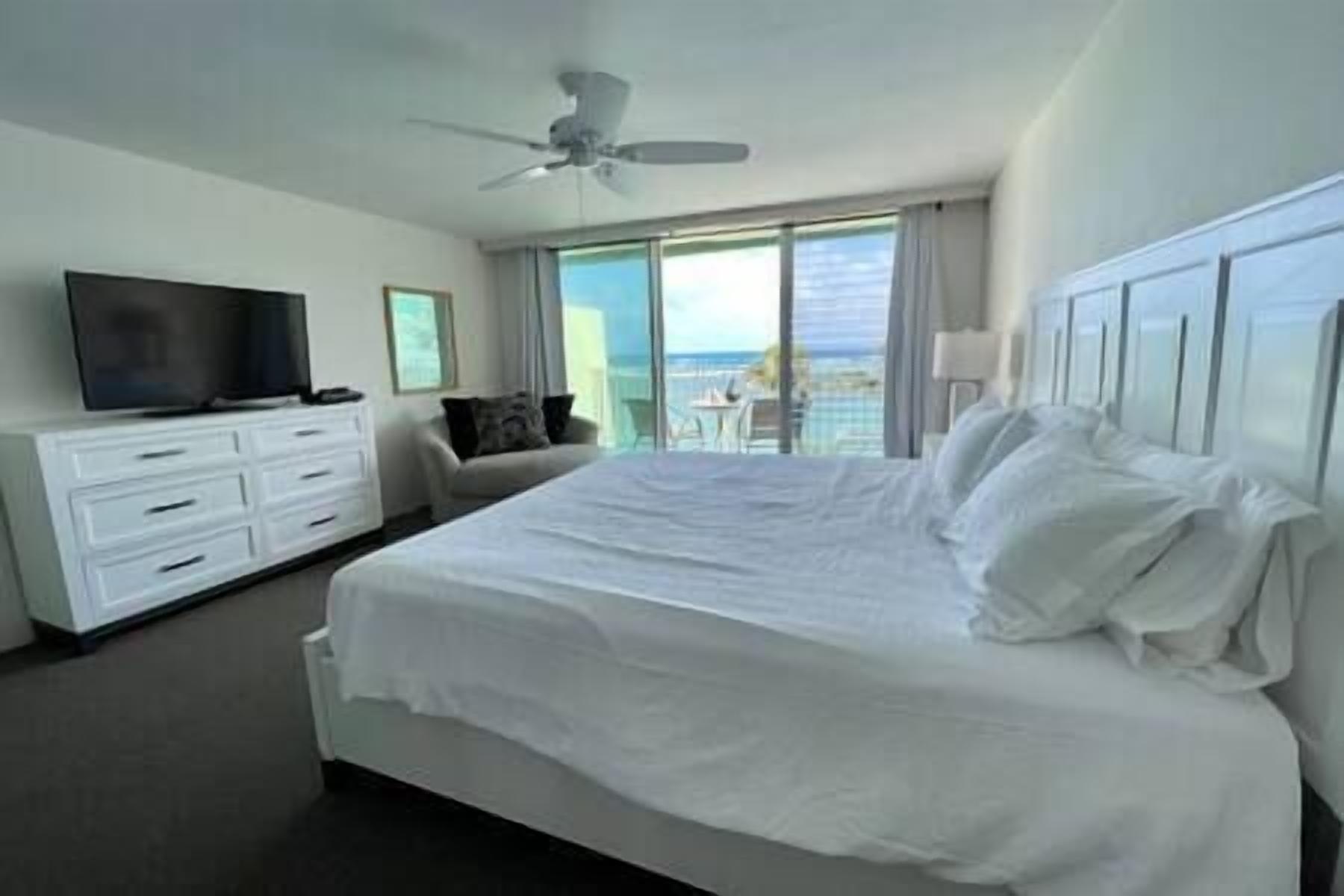 A hotel room with a large bed, a TV, a ceiling fan, and a view of the ocean.