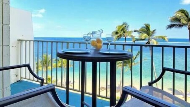 A balcony with a table set for two, overlooking a pool and a beach with palm trees.