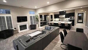 A spacious living area with a large island, a fireplace, and a modern kitchen in the background. - Luxe Desert Oasis: Pool, Spa, Gym & Golf Cart Access Champ (La Quinta)
