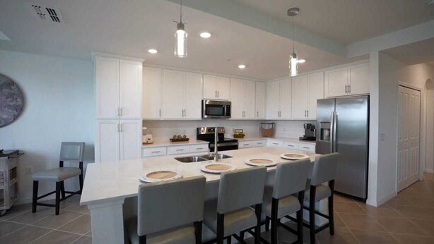 Una cocina moderna con armarios blancos, una isla central con taburetes de bar, electrodomésticos de acero inoxidable e iluminación colgante.