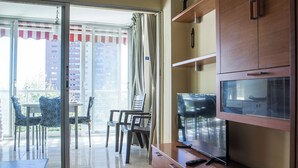 A room with a sliding glass door leading to an outdoor balcony, a television on a wooden stand, and a small table with chairs.