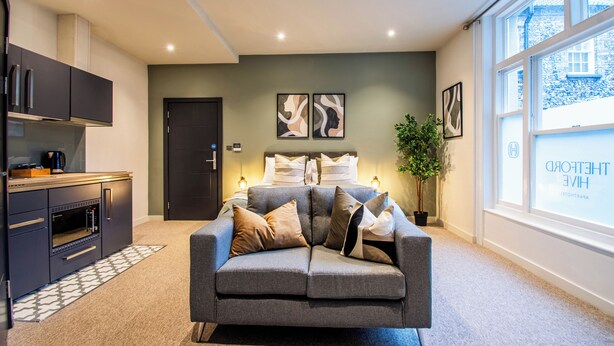 A modern living room with a grey sofa, a kitchen area with dark cabinets, and a large window with a view of a building. - Thetford Hive (Thetford)