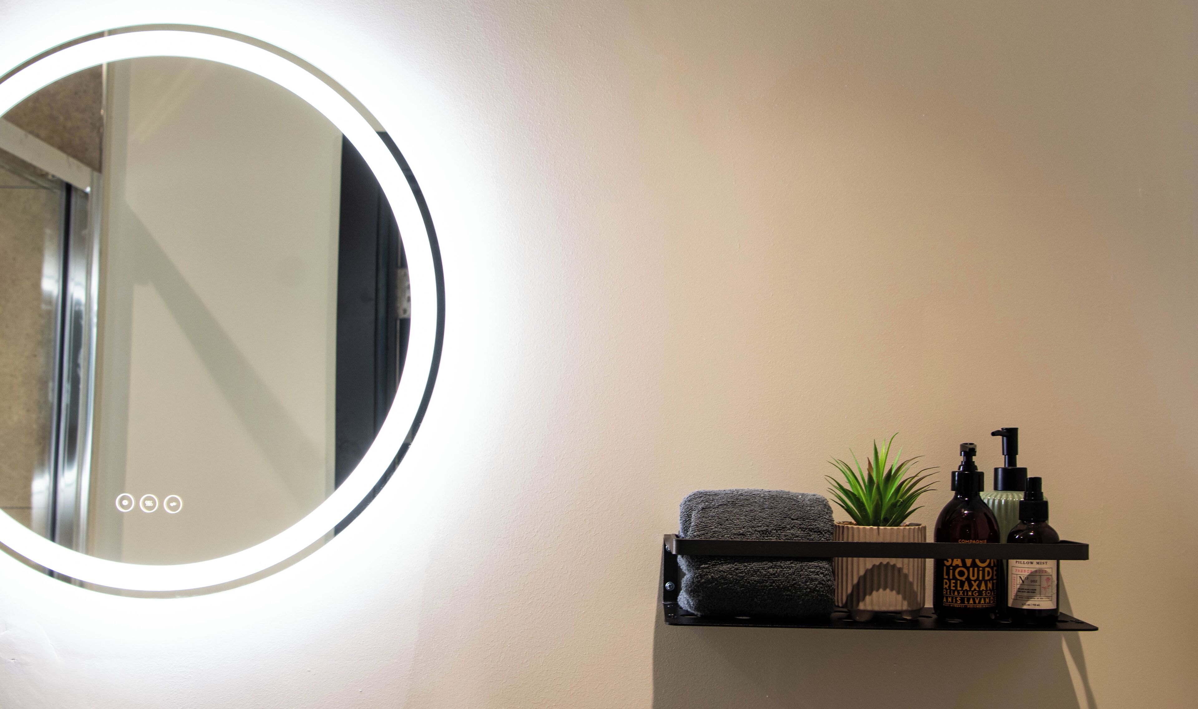 A bathroom with a round mirror, a towel, a plant, and toiletries on a shelf.