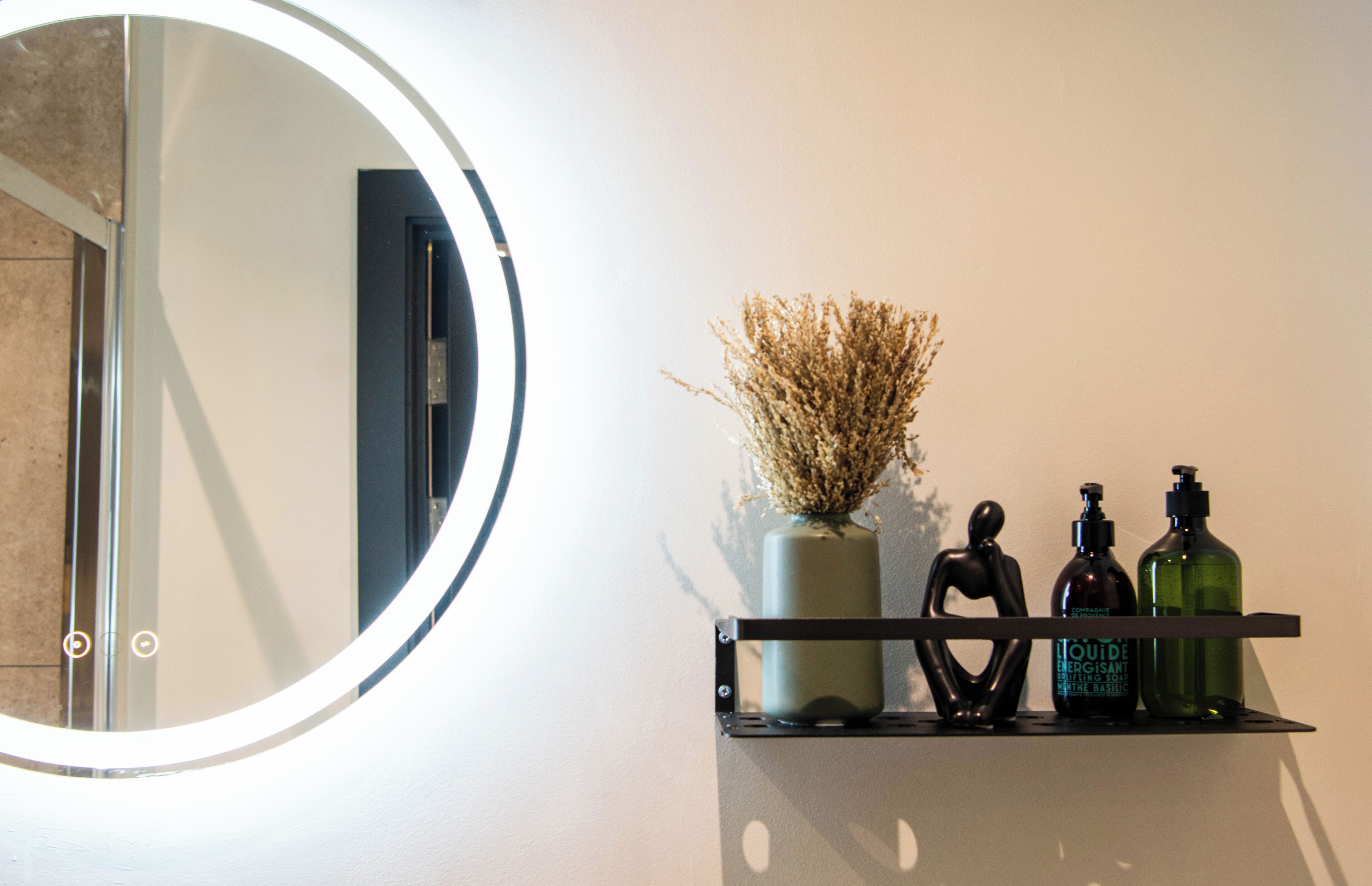 Un bagno con uno specchio rotondo, un vaso con fiori secchi e due flaconi di sapone liquido su uno scaffale.