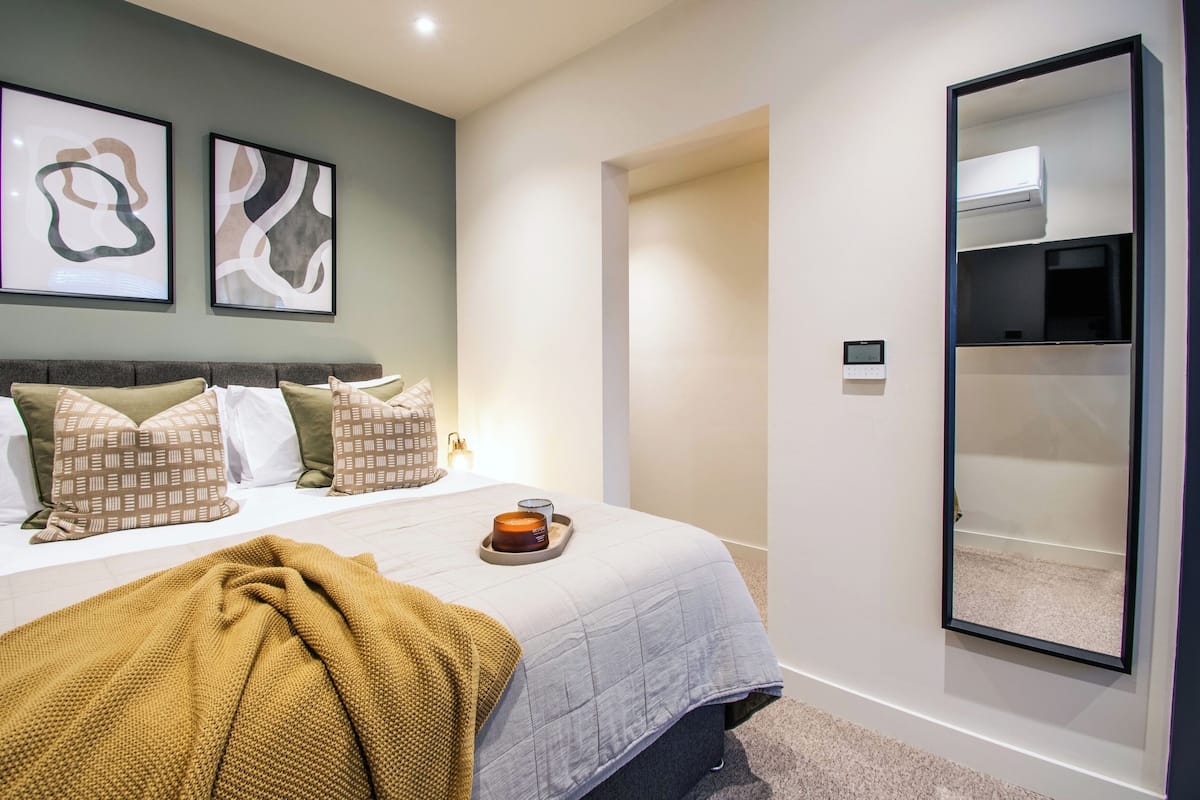 Un dormitorio con cama, almohadas, un edredón, una mesita de noche con una vela y un aire acondicionado instalado en la pared.
