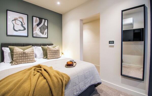 Un dormitorio con cama, almohadas, un edredón, una mesita de noche con una vela y un aire acondicionado instalado en la pared.