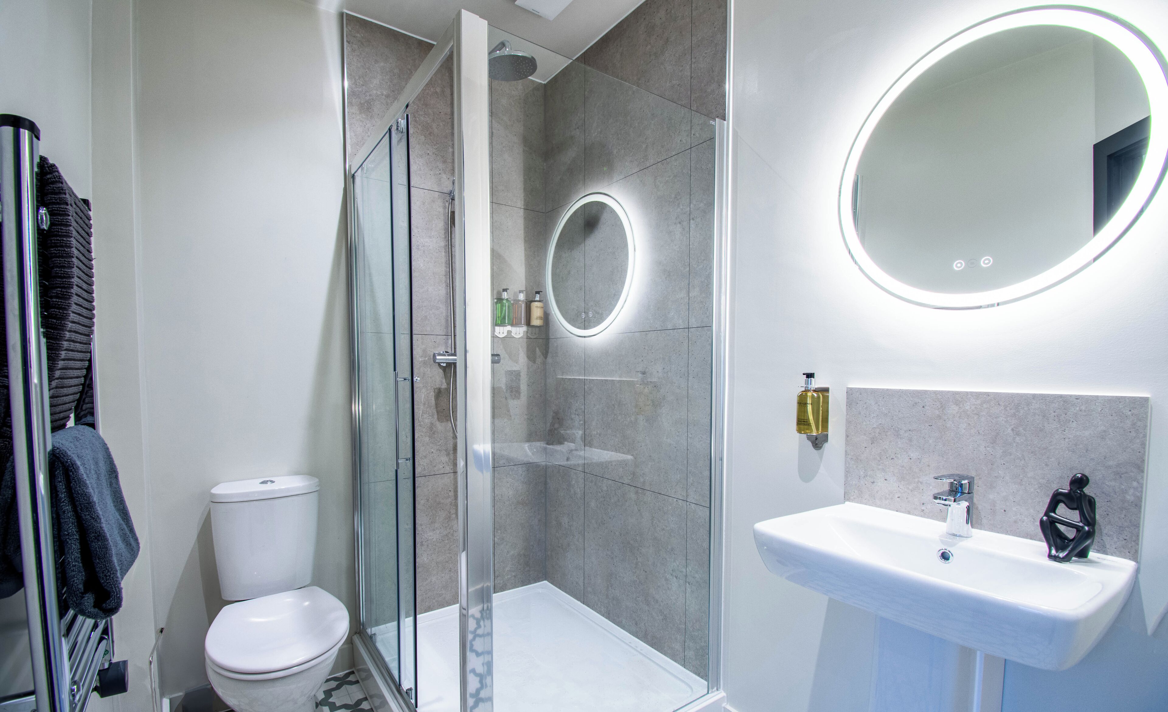 Et moderne badeværelse med et glasafskærmet brusebad, en hvid vask med et rundt spejl og et toilet.