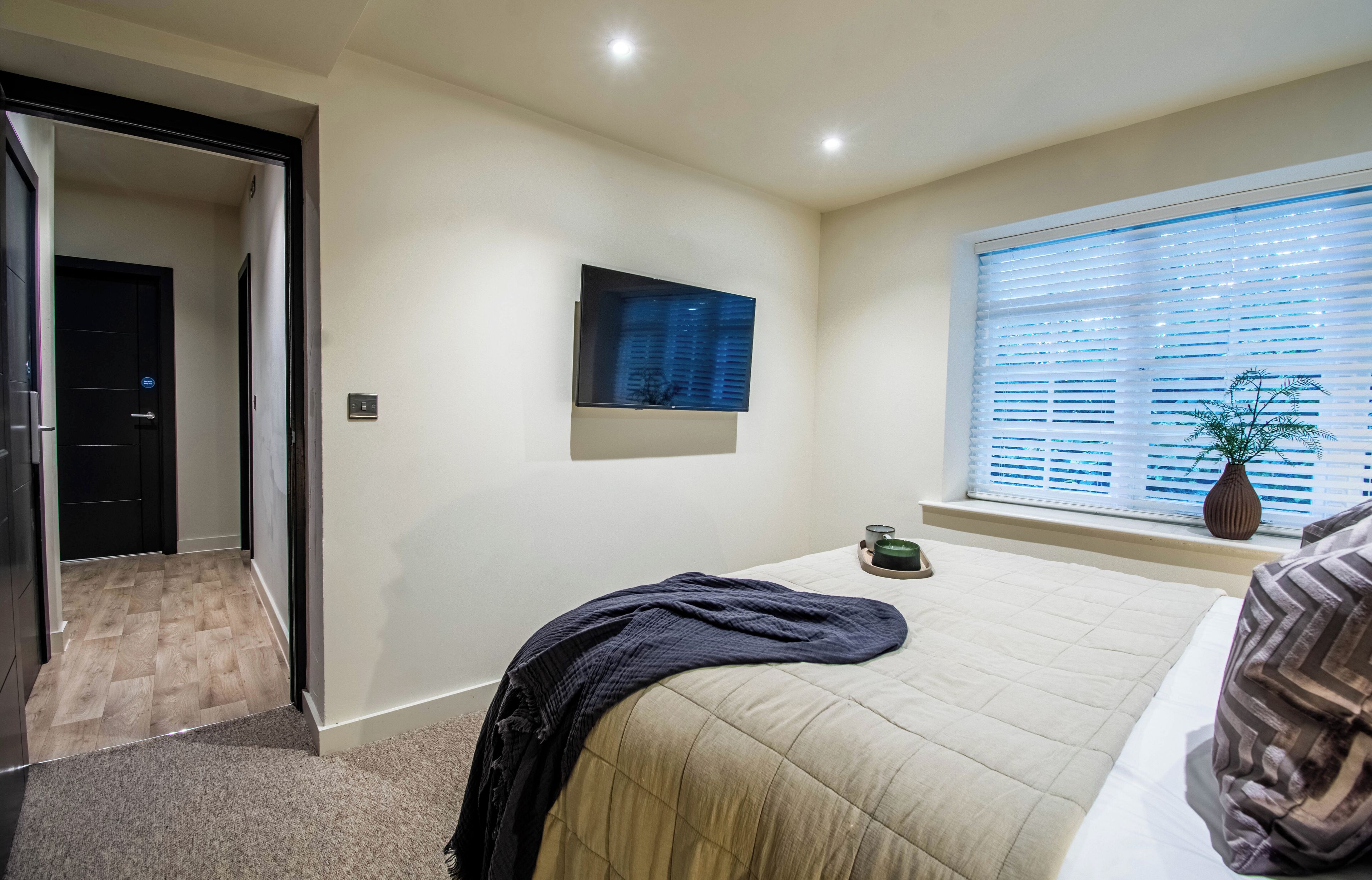Un dormitorio con cama, ventana con persianas, televisor montado en la pared y puerta que da a otra habitación.