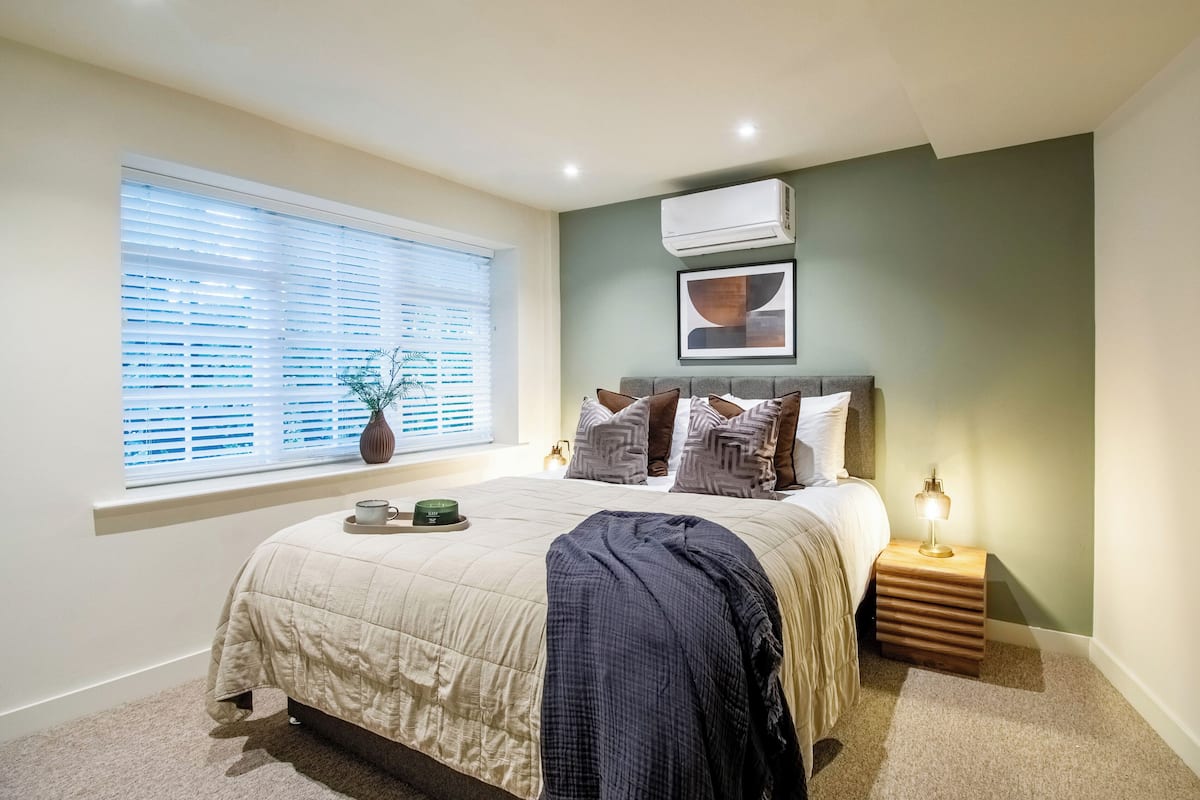 Un dormitorio con cama, mesita de noche, lámpara, ventana con persianas, equipo de aire acondicionado y un cuadro en la pared.