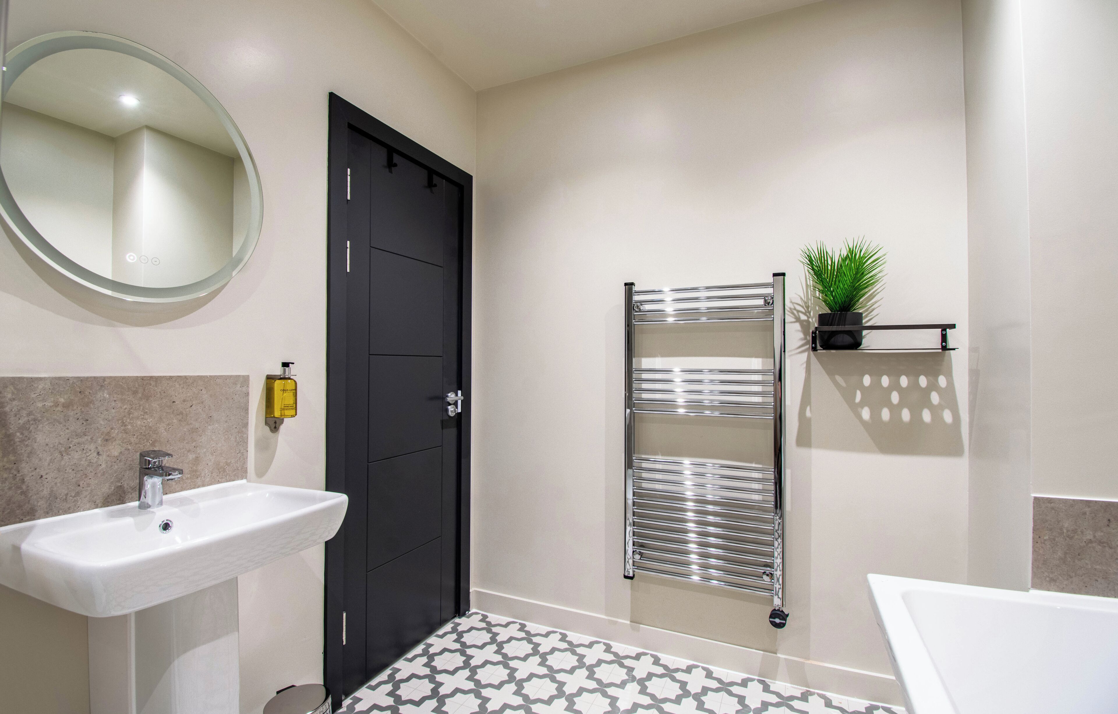 Un baño moderno con un lavabo blanco, una puerta negra, un perchero con una planta y un suelo estampado.