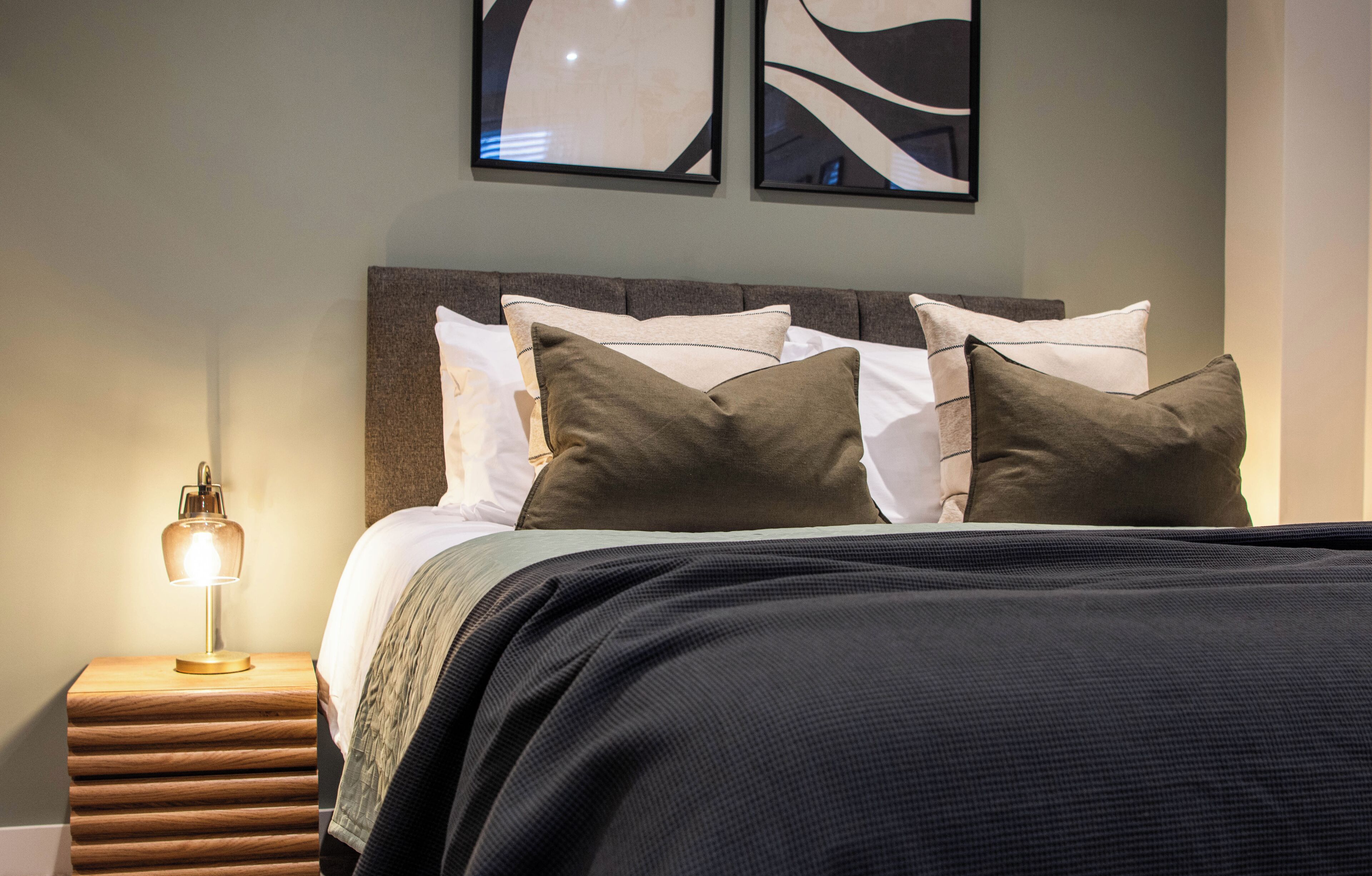Un lit bien fait, recouvert d’une couvre-lit sombre et agrémenté de plusieurs oreillers, une lampe de chevet et une œuvre d’art abstraite accrochée au mur au-dessus du lit.