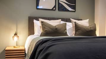 Un lit bien fait, recouvert d’une couvre-lit sombre et agrémenté de plusieurs oreillers, une lampe de chevet et une œuvre d’art abstraite accrochée au mur au-dessus du lit.