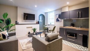 A modern living room with a sofa, coffee table, and a TV mounted on the wall. There is a small dining area with a table and chairs, and a kitchen area with a microwave and oven. - Thetford Hive (Thetford)