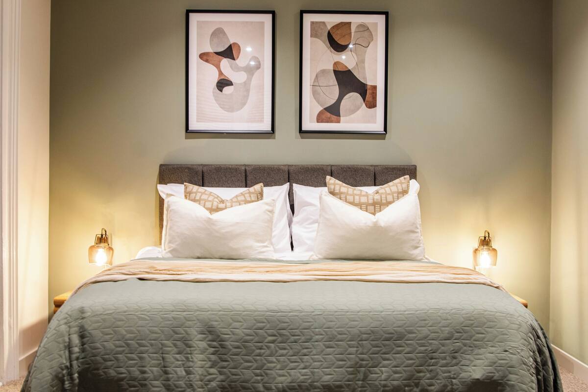 Una cama bien tendida con una colcha verde acolchada, dos cuadros abstractos enmarcados encima y dos lámparas de noche a cada lado.