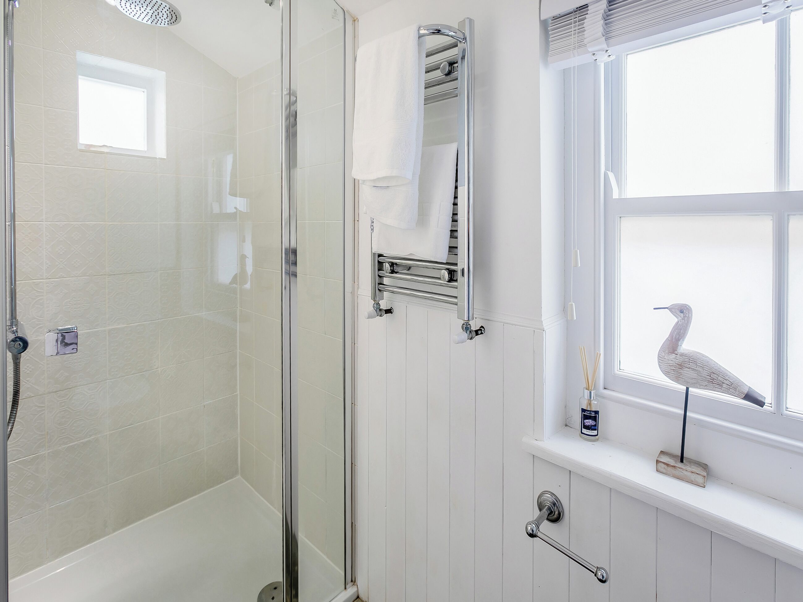 A bathroom with a glass-enclosed shower, a white tiled wall, a towel rack, and a window with a bird outside.