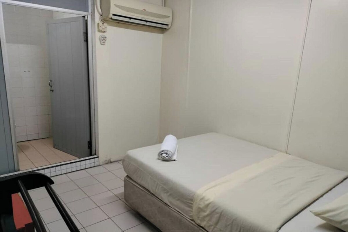 Een kleine kamer met een enkel bed, een witte matras, een opgerolde handdoek op het bed, een wandairconditioner en een deur naar een badkamer.