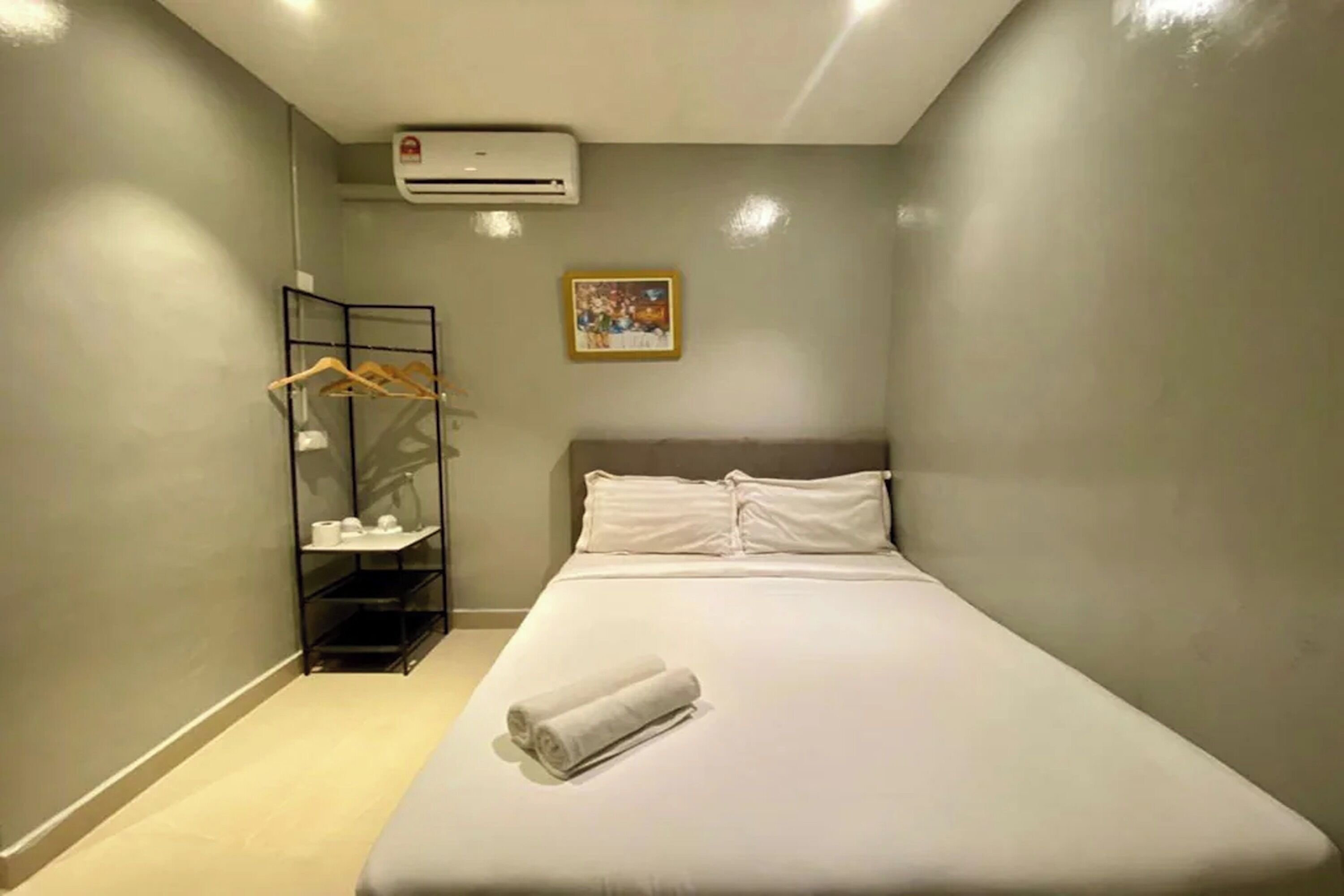 Une chambre d’hôtel avec un lit, un climatiseur fixé au mur, un tableau encadré et une étagère contenant des articles de toilette.