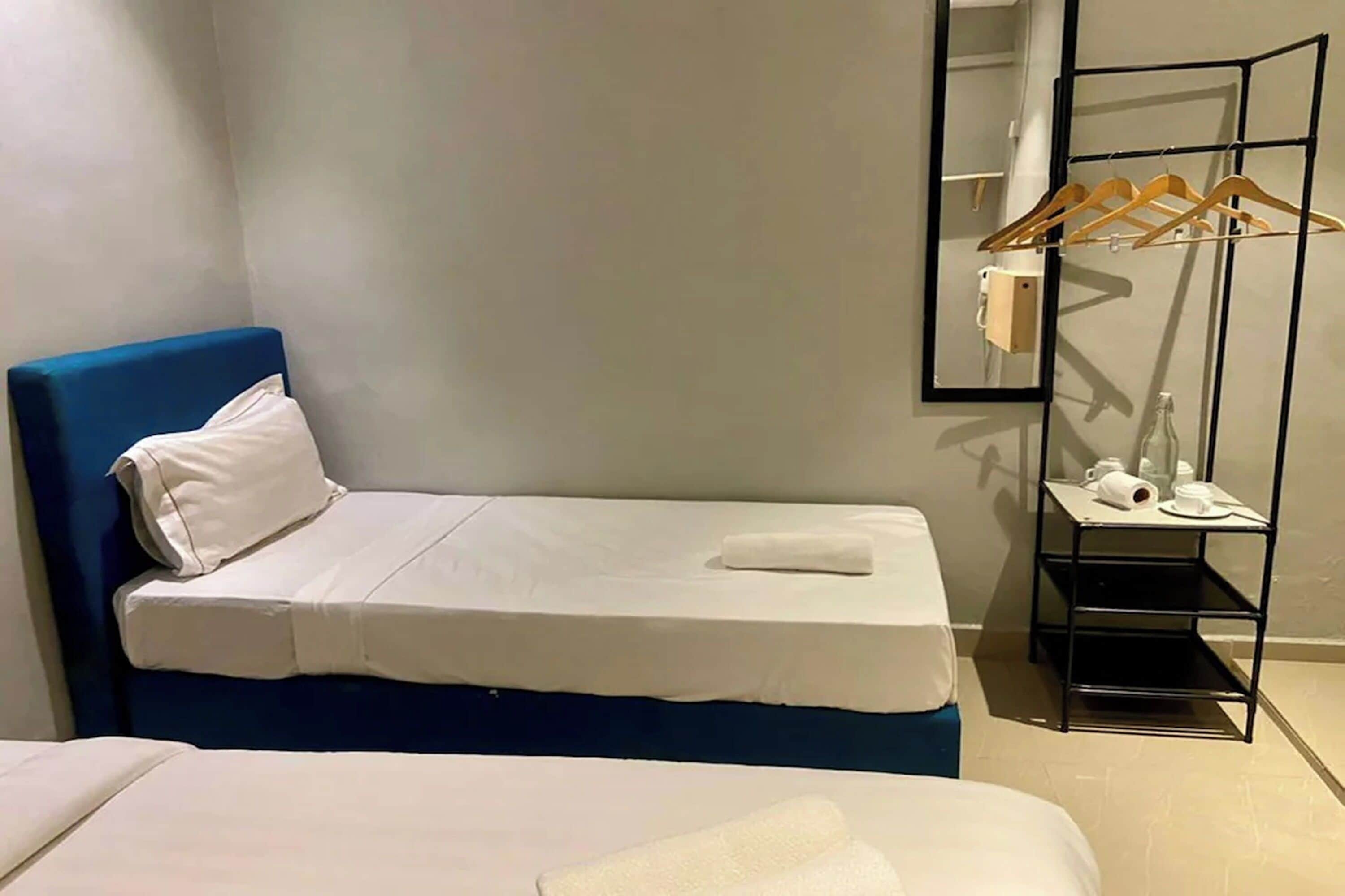 A small, neatly arranged bedroom with a single bed, a bedside table with a cup and a bottle, and a wall-mounted shelf with hanging clothes.
